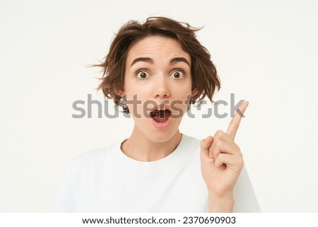 Excited girl has an idea, thinks of solution, pointing one finger and smiling, standing over white background.