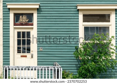 The exterior of a green colored country style wooden house with a half glass door. There's a letter slot in the door and a small model boat in the window. The building has a closed double hung window.