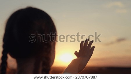 Cute little girl reaches hand to sunset sun disk making wish in evening field