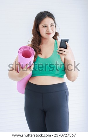 Asia young chubby fat healthy oversized overweight plus size female sportswoman in sportswear sports bra legging sneakers standing holding yoga pilates mat looking at water bottle in home fitness gym. Royalty-Free Stock Photo #2370727367