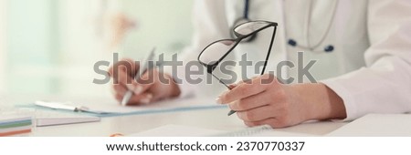 Female doctor writes notes holding glasses at table in hospital office closeup. Therapist fills medical records of patient at workplace