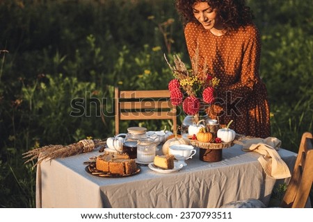 Romantic atmosphere, homemade pie, pumpkins as decor. Sunset, golden hour. Beautiful young woman in a long dress arranging dinner table for a wedding or thanksgiving celebration outdoors, in a meadow Royalty-Free Stock Photo #2370793511