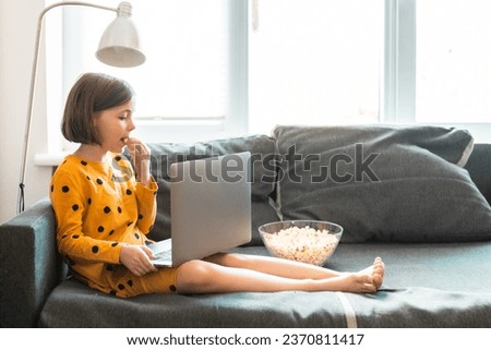 Little girl with laptop and popsorn sits on a grey couach at the bookshelves. School girl watches educational video. children and cartoons concept.