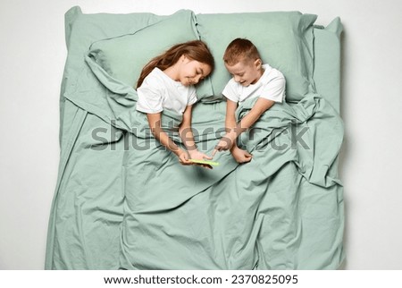 Top view portrait of little girl and boy lying on back covered with blanket looking at phone, watching cartoons. Lovely kid, child in big mint bed. Concept of emotions, sleep wellness, awaking. Ad