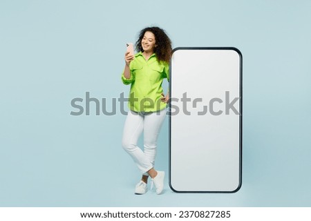 Full body young latin woman wear green shirt casual clothes big huge blank screen mobile cell phone smartphone with workspace area use mobile cell phone isolated on plain light blue cyan background