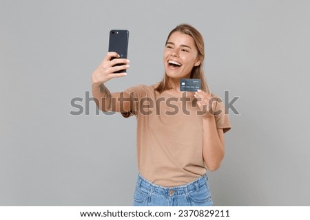 Cheerful young blonde woman in casual beige t-shirt posing isolated on gray background studio. People lifestyle concept. Mock up copy space. Doing selfie shot on mobile phone hold credit bank card