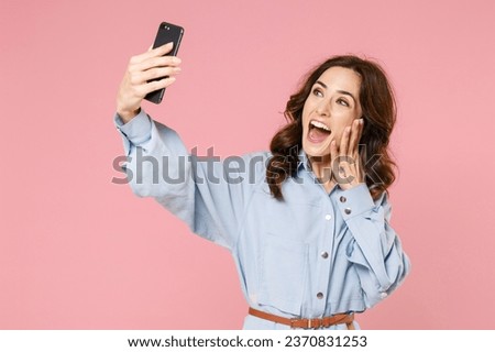 Excited cheerful young brunette woman 20s wearing casual blue shirt dress posing standing doing selfie shot on mobile phone put hand on cheek isolated on pastel pink colour background studio portrait