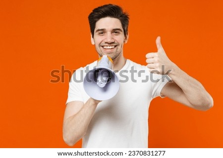Smiling funny young man 20s wearing basic casual empty blank white t-shirt standing screaming in megaphone showing thumb up looking camera isolated on bright orange colour background studio portrait