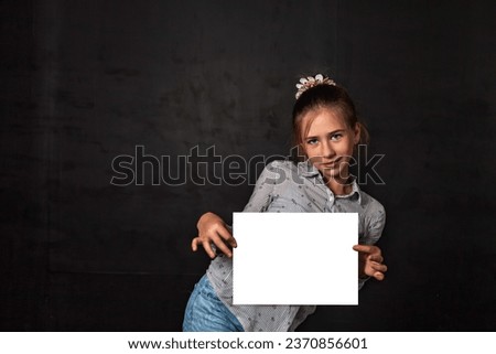 Banner of cute child girl holding white empty blank frame, posing at black backdrop, smile looking at camera. Kid promote mockup template, blank paper in hands. Advertising concept. Copy ad text space