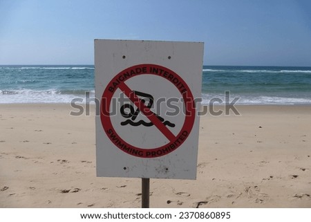 On a French beach, a sign displays the words "No Swimming" in bold letters. Waves lap gently against the shore.