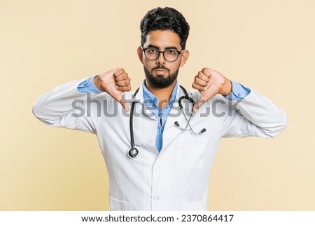 Dislike. Upset unhappy Indian young doctor cardiologist man showing thumbs down sign gesture, disapproval, dissatisfied, negative feedback. Arabian apothecary pharmacy guy isolated on beige background Royalty-Free Stock Photo #2370864417