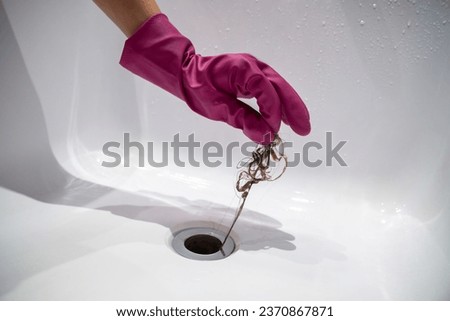 Hand of woman in pink rubber glove taking of hair clump from water drain, blocked pipe in bath. Woman cleaning sewer trap. Bath plug hole blocked with dirt. Problem with shower sewerage in bathroom. Royalty-Free Stock Photo #2370867871
