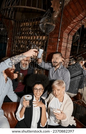 cheerful multicultural women looking at camera near bearded colleagues toasting in cocktail bar