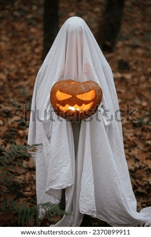 Spooky ghost holding glowing jack o lantern in moody dark autumn forest. Person dressed in white sheet as ghost with pumpkin standing in evening fall woods. Happy Halloween! Trick or treat