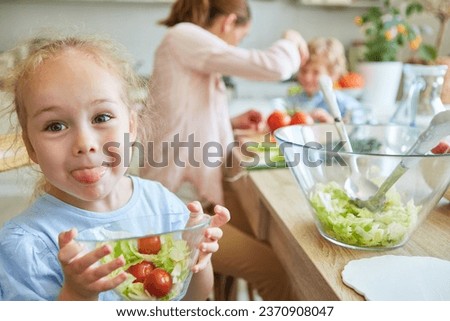 Playful girl sticking out tongue while holding salad bowl in kitchen at home