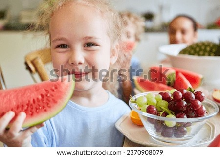 Cute blond girl smiling while holding watermelon slice with grapes in bowl at home