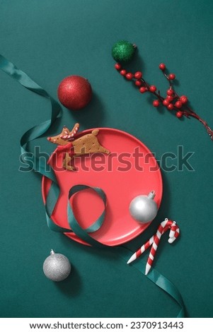 Top view of a red round plate displayed with decoration such as: baubles, candy canes, ribbon and cute reindeer on a green background. Christmas holiday concept and space for design
