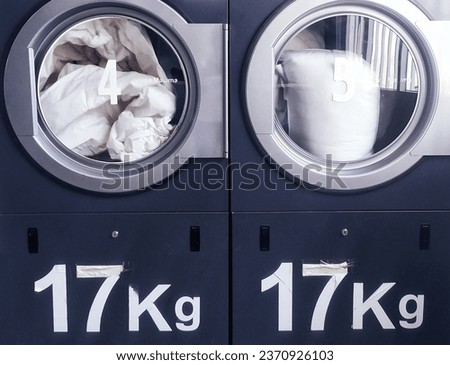 Explore the modern, cosmopolitan lifestyle of shared laundry spaces with this abstract photograph. In the foreground, two well-worn but functional dryers operate with white laundry inside.