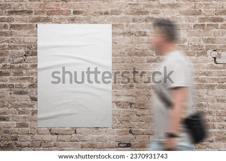 Wrinkled, blank A3 poster in white, adhered to a brick wall. A man walks beside. Ad or marketing campaign design promotion Royalty-Free Stock Photo #2370931743