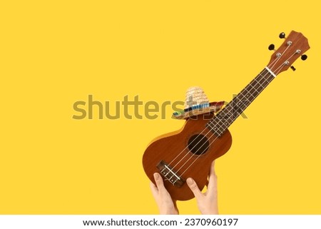 Female hands holding ukulele with sombrero on yellow background Royalty-Free Stock Photo #2370960197