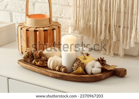Wooden board with burning candles and autumn decor on chest of drawers in room