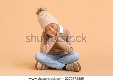 Cute African-American boy in warm winter clothes sitting against beige background