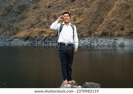 Boy with white shirt and a backpack standing by the lake in the mountains chatting on the phone ,lifestyle Royalty-Free Stock Photo #2370982119