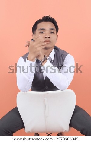 Young Asian businessman wearing a suit vest stares at the camera sitting on a white chair seen from behind with confidence