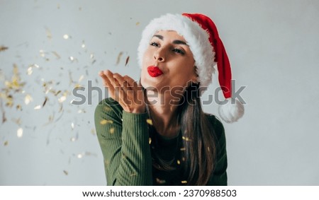 Close up photo of excited young cute woman in Christmas hat is blowing confetti from her hands and celebrating New Year on party