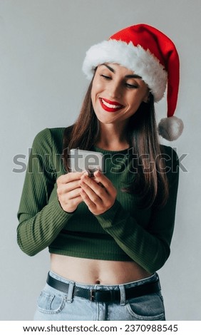 Close up photo of cute smiling woman in Santa Claus hat texting and surfing in net with help of mobile cell smart phone isolated on gray background studio. Happy New Year 2024. Online shopping
