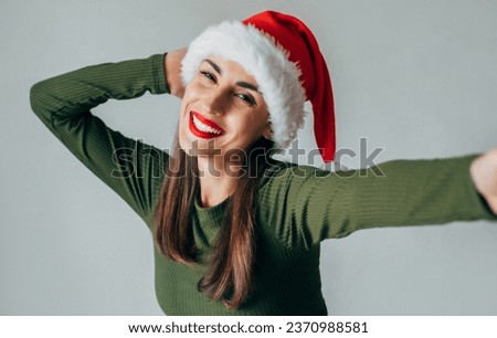 Close up portrait of a pretty cute young woman wearing Christmas Santa hat and taking a selfie isolated over gray background. Happy New Year 2024 and Happy Christmas.