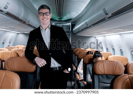 Portrait of happy smiling businessman in black suit, standing on aisle inside airplane, male passenger traveling on business trip by aircraft, businesspeople traveling with airline transportation. Royalty-Free Stock Photo #2370997905