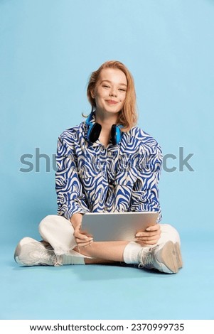Cute smiling young girl, student sitting on floor with tablet over blue studio background. Education, communication, hobby and freelance job. Concept of youth, emotions, lifestyle, facial expression