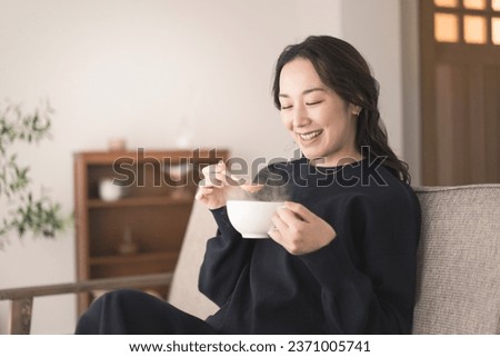 An Asian woman is sitting on the sofa, holding a cup and drinking soup. She is wearing long sleeves. Royalty-Free Stock Photo #2371005741