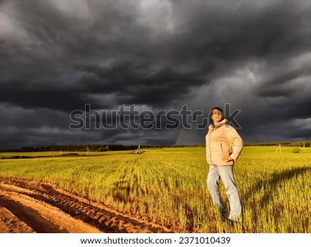 An adult girl in a field and with a stormy sky with clouds posing for picture in the rain. A woman having fun outdoors on rural and rustic nature