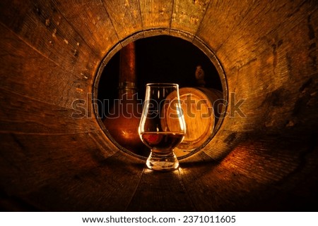 A glass of scotch whiskey in old oak barrel. Copper alambic and small barrel on background. Traditional alcohol distillery concept Royalty-Free Stock Photo #2371011605