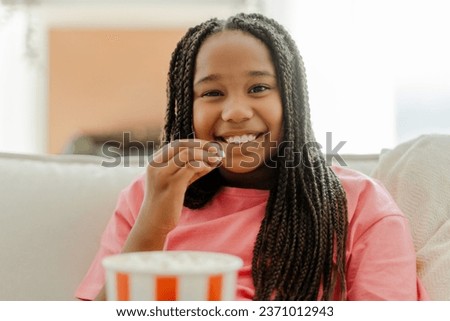 Happy African American girl eating popcorn while watching TV in living room at home. Entertainment, movie and cartoons concept