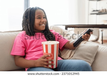 Happy little girl using remote control, eating popcorn and watching tv in living room interior. Entertainment, movie and cartoons concept
