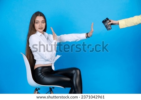 Pretty girl making stop gesture for crop anonymous hand offering beverage. Side view of serious woman outstretching arm in refusion, while looking at camera, isolated on blue. Concept of gesturing. Royalty-Free Stock Photo #2371042961