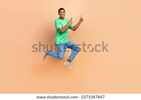 Full size photo of positive guy dressed green t-shirt denim pants jumping directing at sale empty space isolated on beige color background