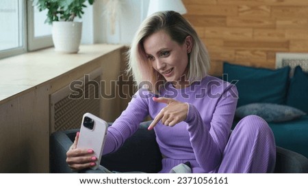 Happy young woman 20 years old in casual clothes, purple home suit sitting on a sofa indoors, relaxing at home in her own apartment room. The blonde girl is holding a smartphone in her hands and