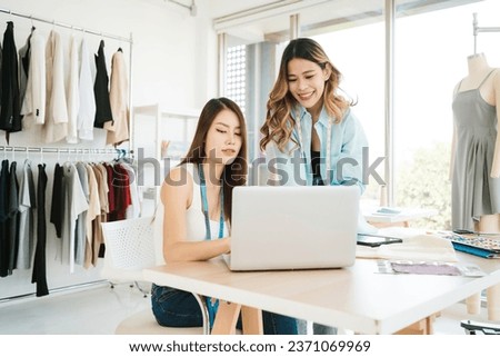 Two Asian female fashion designers or dressmakers brainstormed and discuss the design of the new collection with examples of fabrics and sketches on the laptop. Working together in the studio.
