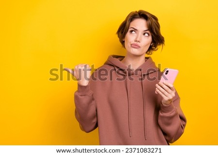 Portrait of funny minded ponder girl wear brown sportswear look directing empty space hold smartphone isolated on yellow color background