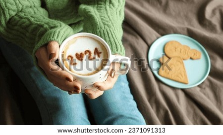 New year 2024 celebrated coffee cup with number 2024 on frothy surface of cappuccino in coffee cup holding by woman in green knitted sweater with jeans sitting on brown bed with gingerbread cookies. Royalty-Free Stock Photo #2371091313