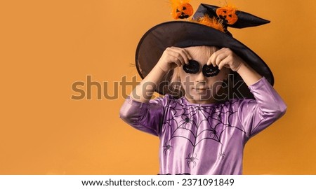 Little girl wearing violet Halloween dress and witch hat covered with spider net with black spooky spiders on it. Orange background. Happy Halloween concept
