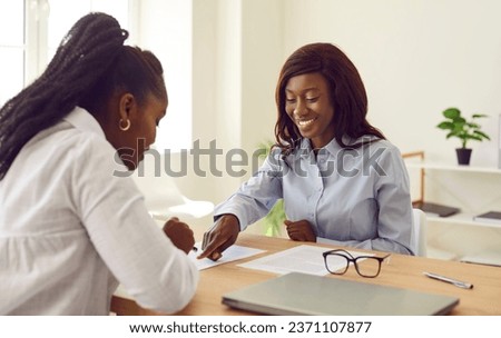 People during meeting in office sign paper document confirming closing of deal. Smiling African American woman showing her female client place to sign contract. Business partnership concept.