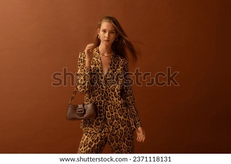 Fashionable confident woman wearing trendy leopard print satin suit blazer, trousers, holding small leather bag, posing in studio, on brown background. Copy, empty space for text