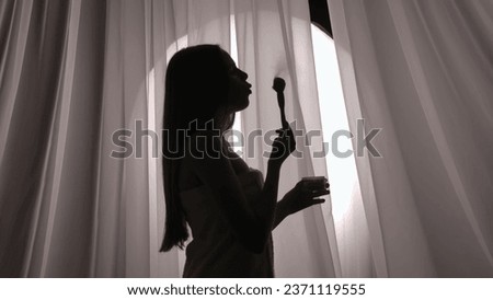 Medium side view shot of a young woman silhouette wrapped in a towel holding a makeup powder jar, blowing on a brush with a product in a muffled light. Royalty-Free Stock Photo #2371119555