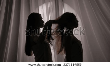 Medium side view shot of two sisters, friends, young women's silhouettes wrapped in a towel. One of them is braiding the other one's hair in a muffled light. Royalty-Free Stock Photo #2371119559
