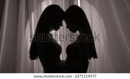 Medium side view shot of two seminaked sisters, young women's silhouettes. They are leaning their foreheads against each other in a muffled light. Royalty-Free Stock Photo #2371119577